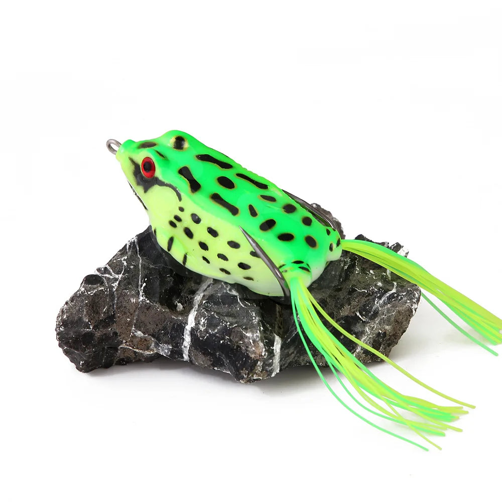 Green frog-shaped fishing lure with black spots and red eyes on a rock, outdoor gear accessory