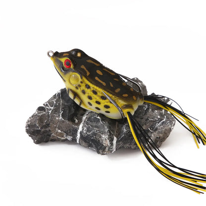 Realistic frog fishing lure with yellow spots, red eyes, and skirted tail on rock, outdoor gear