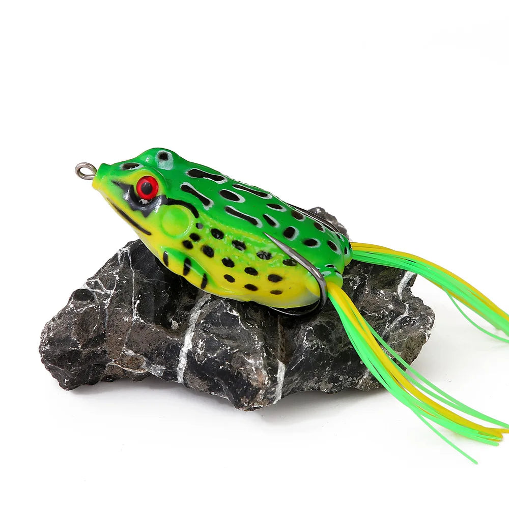 Green frog-shaped fishing lure with red eyes and yellow accents on a rock, outdoor gear