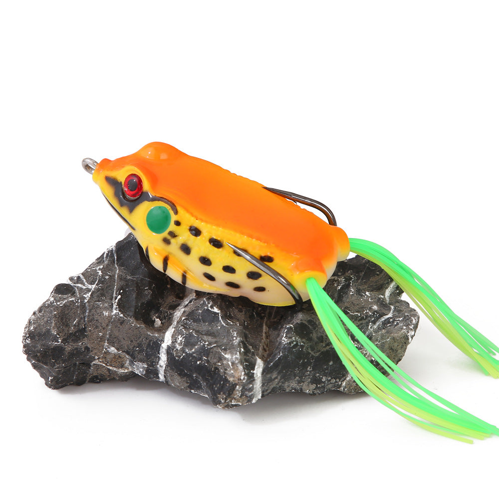 Bright orange and yellow frog fishing lure with green rubber legs on a dark rock, outdoor gear