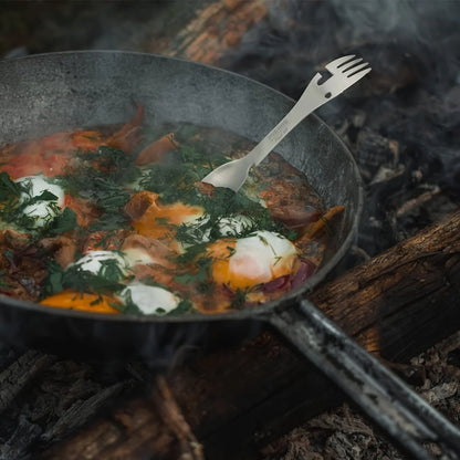Stainless steel 5-in-1 survival tool pitchfork style in a cooking firepot with meat and herbs outdoors.